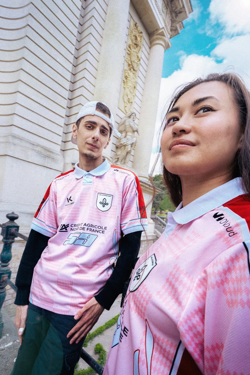 Deux membres de Lille Esport portent le maillot rose de la saison 2025, devant une façade à colonnes sous un ciel bleu.